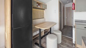 A compact kitchenette with a black refrigerator, a wooden table, two stools, and a small shelf with glassware. - Brand New Luxury Silver Caravan (Ingoldmells, Skegness)