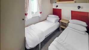 A small, compact room with two single beds, a small table, and a window with curtains. - Brand New Luxury Silver Caravan (Ingoldmells, Skegness)