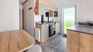 A modern kitchen with a built-in oven, microwave, and kettle, a wooden dining table, and a view of the outdoors. - Brand New Luxury Silver Caravan (Ingoldmells, Skegness)