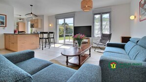 Ein modernes Wohnzimmer mit einer Couch, einem Couchtisch und einem Essbereich mit Stühlen. Es gibt eine Küche mit Holzausstattung und einem Esstisch. Ein großes Fenster bietet einen Blick ins Freie.