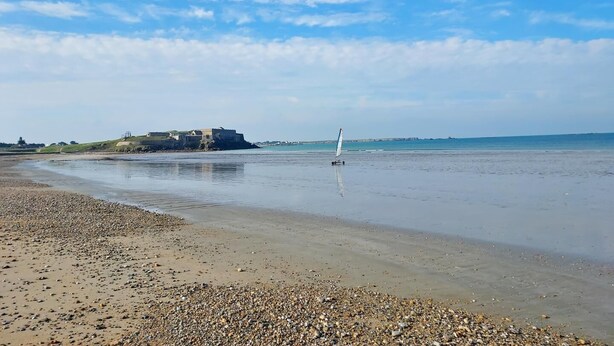 Ein Strand mit Kieselsteinen, ein Segelboot und eine festungsähnliche Struktur im Hintergrund.