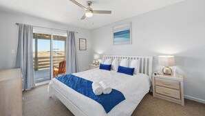 A hotel room with a bed, a ceiling fan, a nightstand with a lamp, a framed picture on the wall, and a view through the window. - Wink Shores | Oceanfront | Private Pool, Hot Tub, and Dog Friendly (Kill Devil Hills)