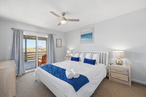 A hotel room with a bed, a ceiling fan, a nightstand with a lamp, a framed picture on the wall, and a view through the window.