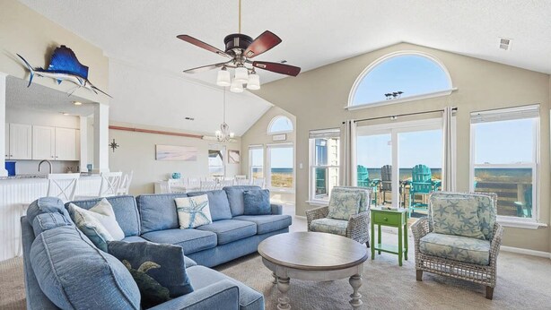 A spacious living room with a blue sofa, a round coffee table, and wicker chairs. A ceiling fan and large windows provide natural light and views of the outdoors. - Wink Shores | Oceanfront | Private Pool, Hot Tub, and Dog Friendly (Kill Devil Hills)