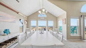 A spacious dining area with a long white table and chairs, a chandelier, and large windows offering a view of the beach. - Wink Shores | Oceanfront | Private Pool, Hot Tub, and Dog Friendly (Kill Devil Hills)