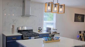 A modern kitchen with a gas stove, a built-in oven, and a coffee machine. There is a window with blinds, a ceiling light fixture, and a painting on the wall. - Cozy Arlington Home • Minutes to AT&T & Globe Life (Arlington)