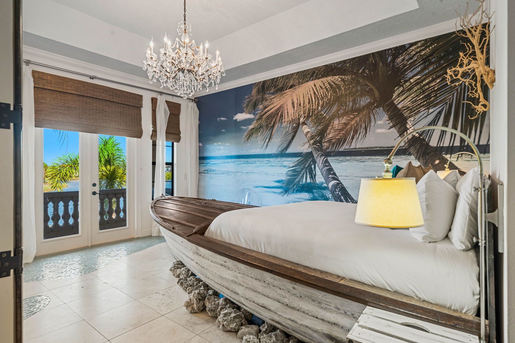 Ein Schlafzimmer mit einem großen Bett, einer Chandelüster, einem Wandbild mit Strandmotiv und Blick aufs Meer durch eine Schiebetür.