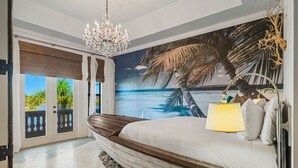 Ein Schlafzimmer mit einem großen Bett, einer Chandelüster, einem Wandbild mit Strandmotiv und Blick aufs Meer durch eine Schiebetür.