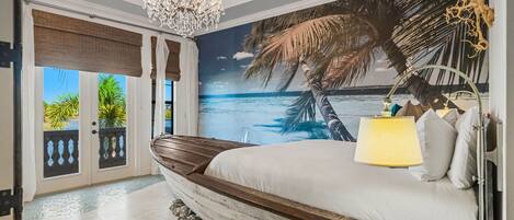 Ein Schlafzimmer mit einem großen Bett, einer Chandelüster, einem Wandbild mit Strandmotiv und Blick aufs Meer durch eine Schiebetür.