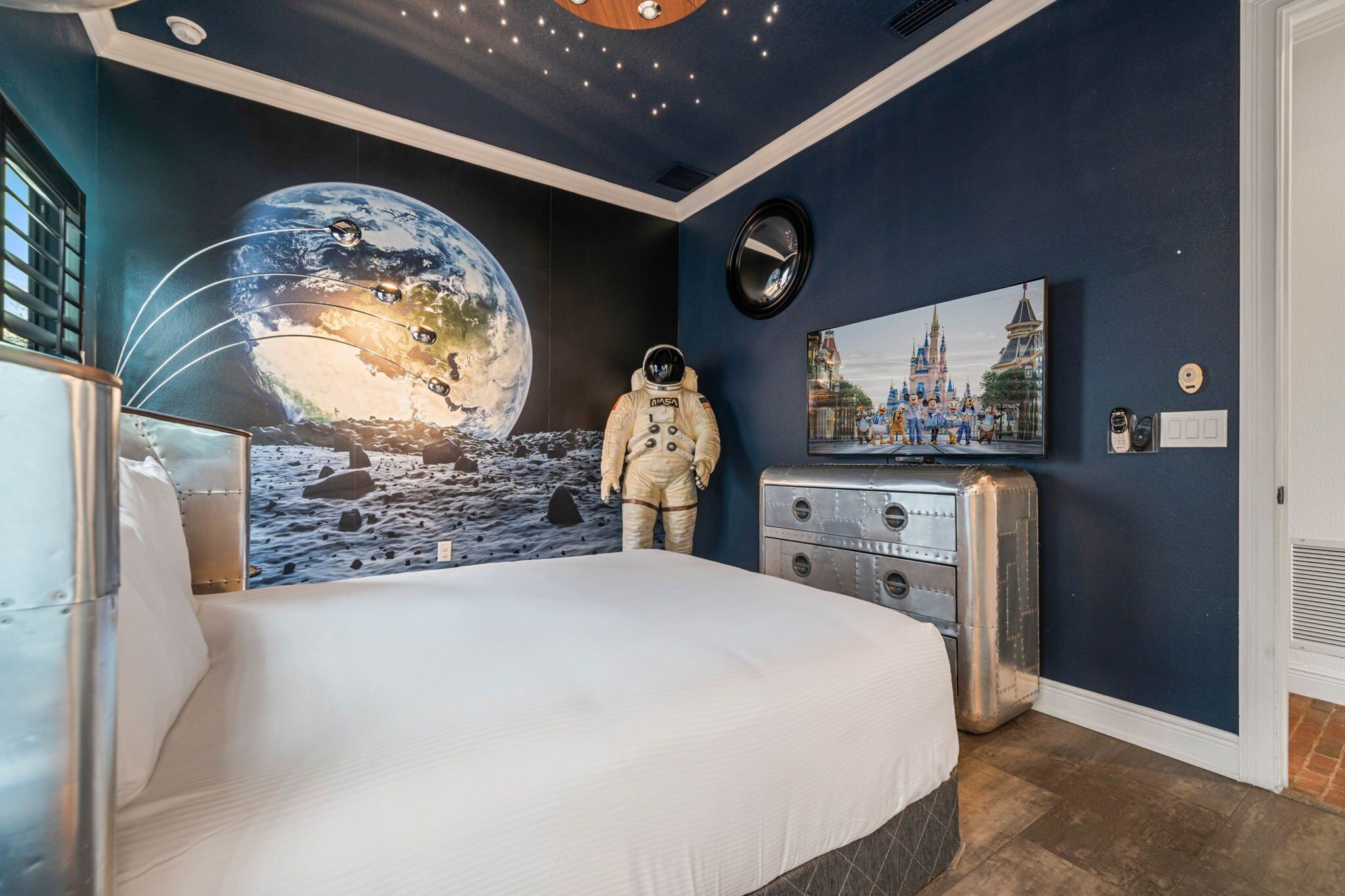A bedroom with a large bed, a mural of Earth, a space suit figure, a vintage radio, and a Disney castle painting.