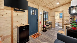 A compact, wood-paneled room with a black TV, a small kitchen area, and a door leading to another room. - Tiny Retreat-Huge Covered Deck,Hot Tub,Lake Access (Sevierville)