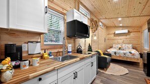 A compact, wooden-floored room with a kitchenette, a bed with a floral bedspread, and a small Christmas tree. - Tiny Retreat-Huge Covered Deck,Hot Tub,Lake Access (Sevierville)