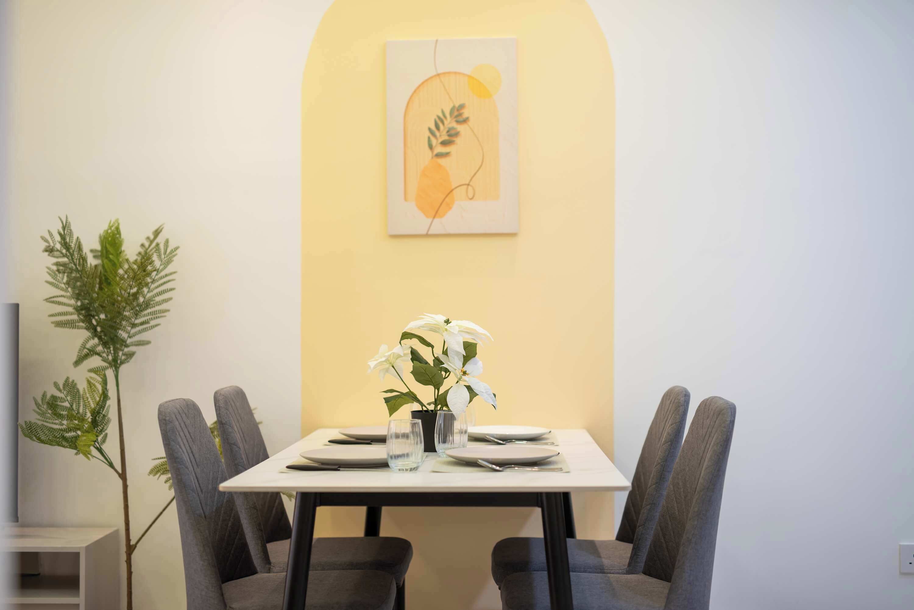 A dining area with a table set for four, a potted plant, and a framed artwork on the wall.