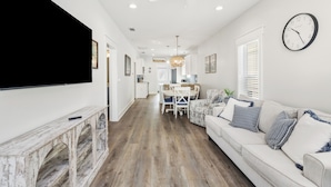 A modern living room with a flat-screen TV, a white sofa with patterned cushions, a wooden console table, and a dining area with a chandelier. - NEW LISTING! Special Sunset is steps to beach, hot tub, fenced backyard and pet friendly! Sleeps 6 (Mexico Beach)