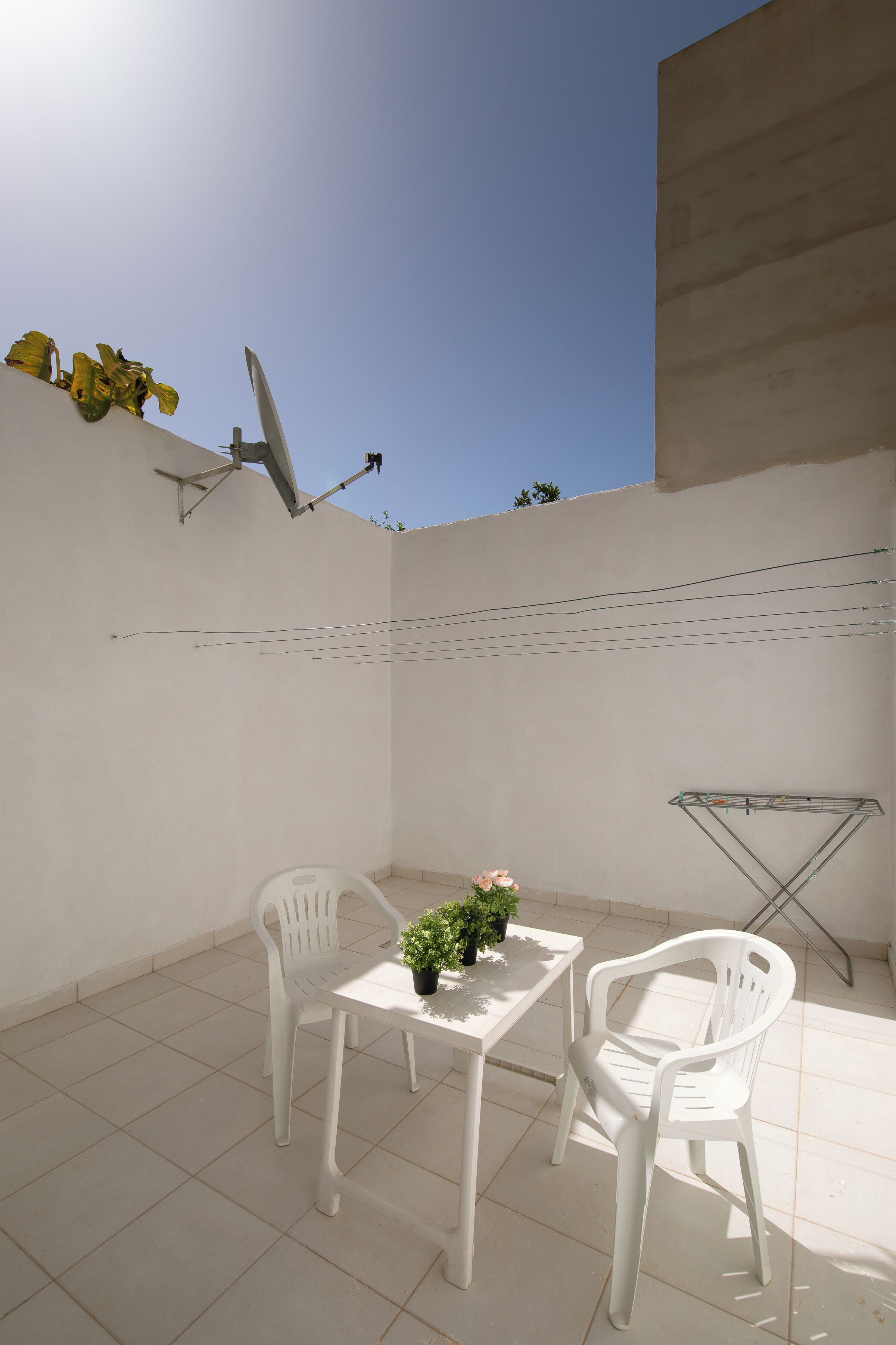 En lille udendørs terrasse med et hvidt bord og to hvide stole, en potteplante og et tørrestativ til tøj.