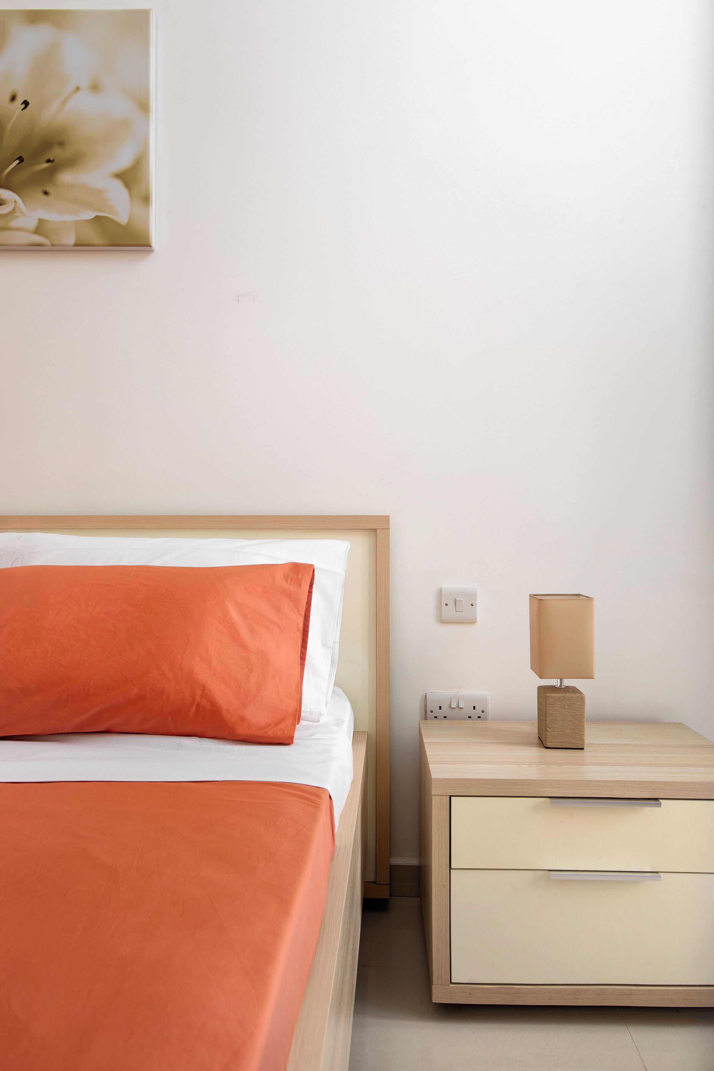 A neatly made bed with an orange pillow, a bedside table with a lamp, and a wall outlet.