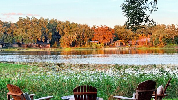 Un patio junto al lago con mecedoras de madera y una mesa, con vista a un lago tranquilo y un área boscosa.