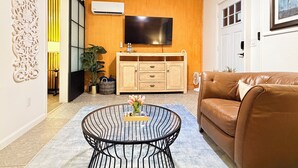 Una sala de estar con un sofá de cuero marrón, una mesa de centro redonda de metal con un jarrón de flores, un mueble de madera con un televisor de pantalla plana y un ventilador de techo.