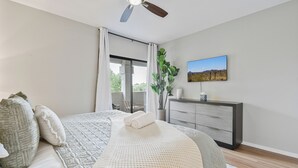 Une chambre à coucher avec un lit, un ventilateur de plafond, une fenêtre avec des rideaux, une plante, un téléviseur et une commode.