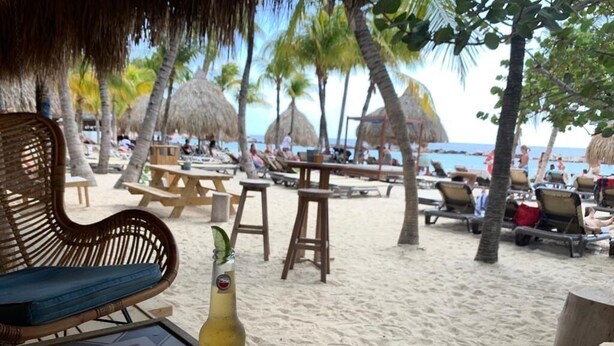 Ein Sitzbereich am Strand mit Holztischen und -stühlen, einem Korbstuhl und einer Bierflasche auf einem Tisch.