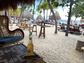 A beachside seating area with wooden tables and chairs, a wicker chair, and a bottle of beer on a table. - Deluxe private Apartment, 90m2 (Jan Thiel)