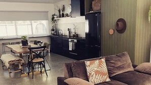 A modern living room with a brown sofa, a dining area with a wooden table and chairs, and a kitchen with dark cabinets and a stainless steel refrigerator. - Premium Halls Gap Holiday Home - Firepit and Local Wine (Halls Gap)