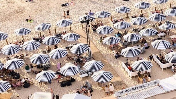 Ein Strand mit mehreren gestreiften Sonnenschirmen und Liegestühlen, der einen Beachclub-Charakter vermittelt.