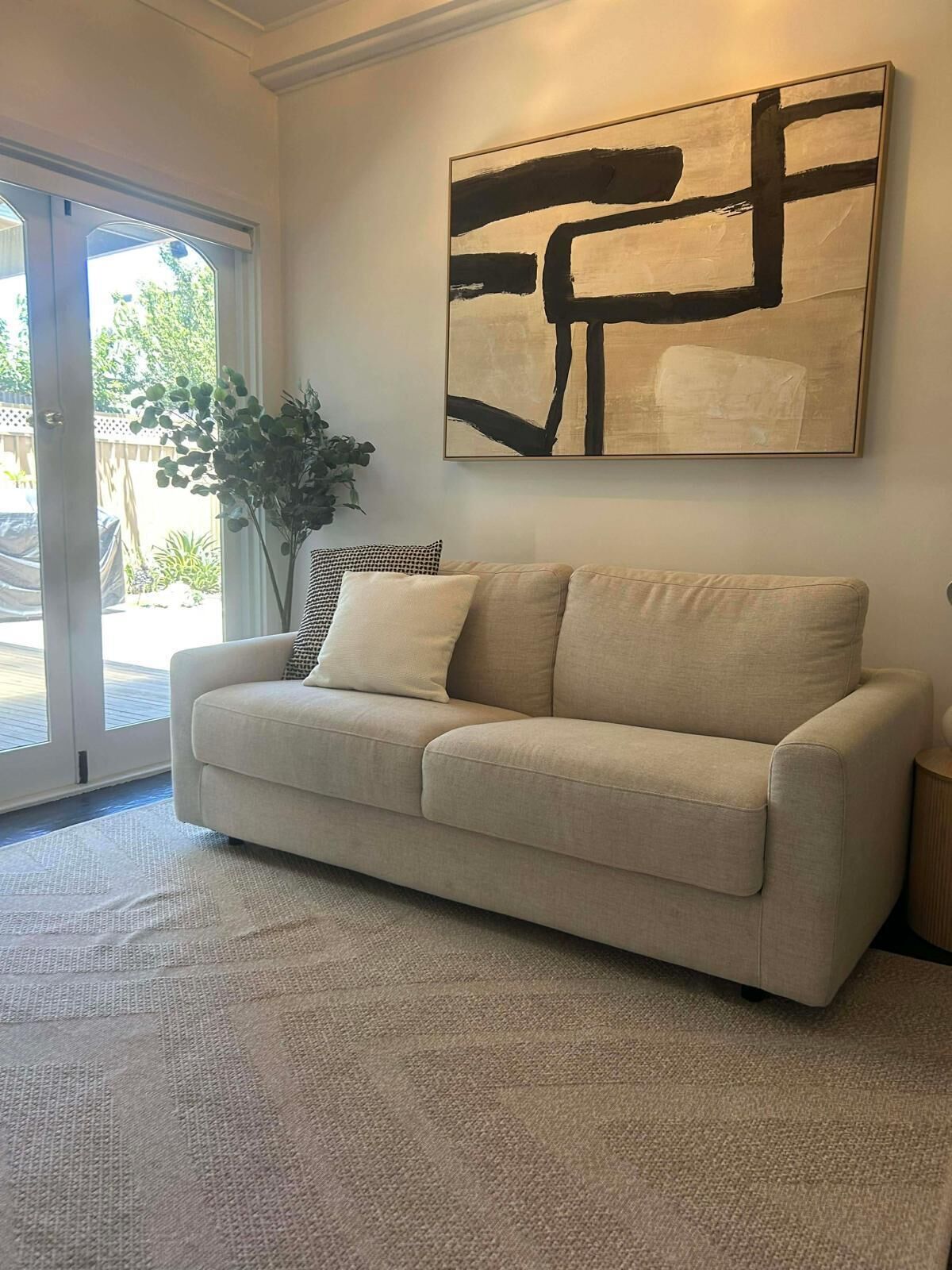 A living room with a beige sofa, a large abstract painting, and a potted plant.