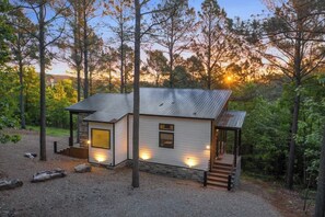 A single-story cabin with a metal roof, surrounded by trees, and featuring a small porch with steps leading to the entrance. - Mini Marshmallow Cabin - Cozy & Romantic GetAway (Broken Bow)
