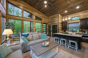 A cabin interior with a wooden ceiling, large windows, a sofa, a coffee table, and a kitchen area. - Mini Marshmallow Cabin - Cozy & Romantic GetAway (Broken Bow)