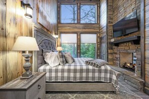 A bedroom with a large bed, a fireplace, a television, and a window with a view of trees. - Mini Marshmallow Cabin - Cozy & Romantic GetAway (Broken Bow)