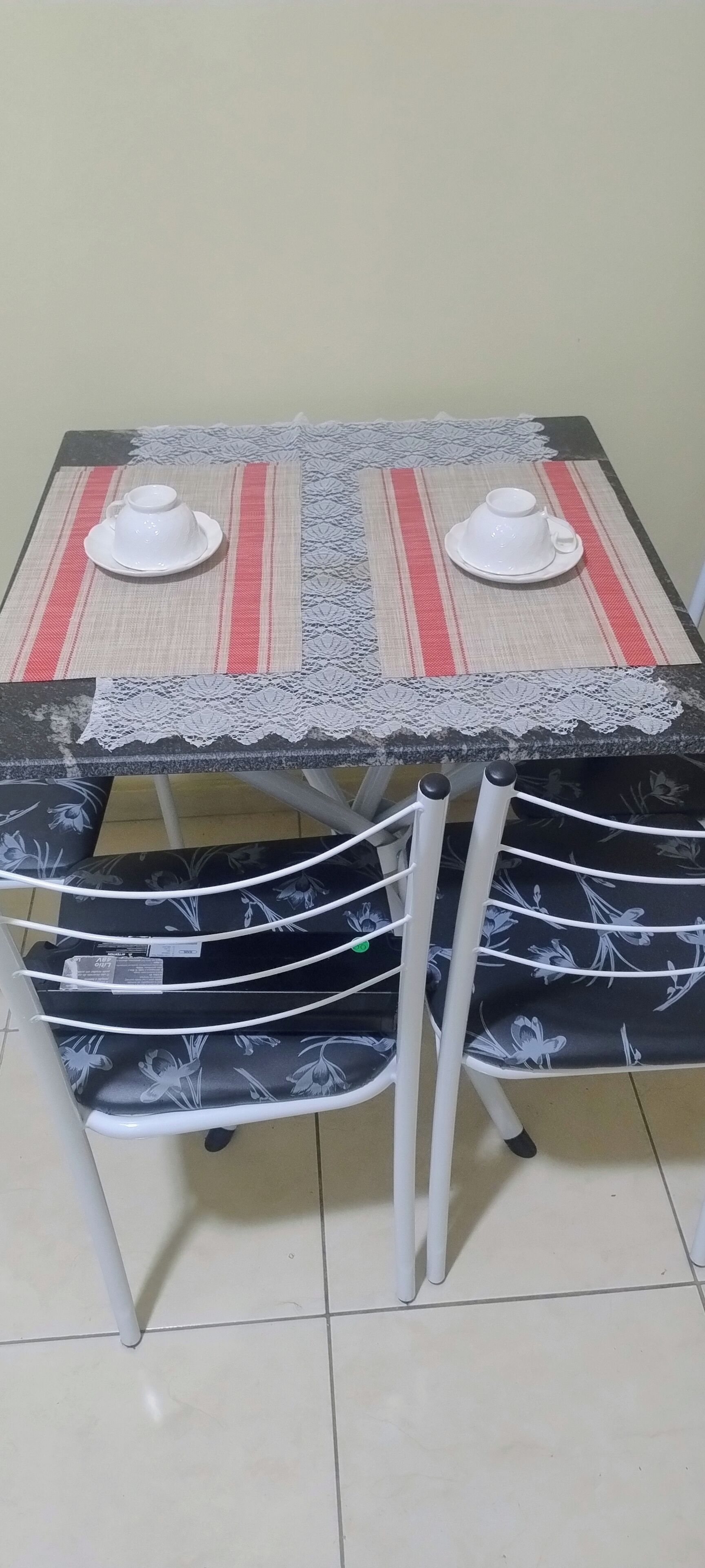 A dining table with two white cups and saucers, a lace table runner, and a chair with a floral pattern.