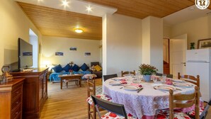 Un comedor con una mesa redonda dispuesta para cuatro personas, rodeada de sillas de madera con tapicería floral. A la izquierda, un aparador de madera con cajones, y un sofá con cojines azules al fondo.