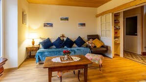 Una sala de estar acogedora con techo de madera, un sofá con cojines azules, un sillón de cuero marrón, una mesa de centro de madera con camino de mesa de encaje, y una mesita con un florero de flores rojas.