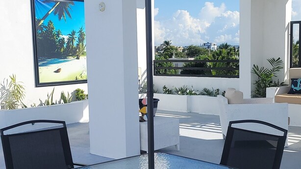 Eine Terrasse im Freien mit einem Glastisch und Stühlen, einem großen Sonnenschirm und Blick auf einen Strand durch ein großes Fenster.