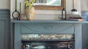 Une cheminée surmontée d’un miroir, deux lampes fixées au mur, un vase avec une plante et une petite pile de livres.