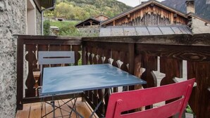 Una terraza de madera con una mesa azul y una silla roja, con vistas a una zona montañosa con edificios tradicionales.