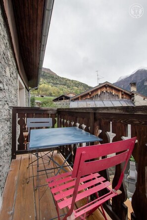 Eine Holzterrasse mit einem blauen Tisch und einem roten Stuhl, mit Blick auf eine gebirgige Landschaft mit traditionellen Gebäuden.