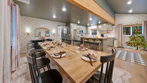 A modern kitchen with white cabinets, a wooden dining table set for four, and a small Christmas tree by the window. - Nostalgic Nook — Offering a gym, sauna, hot tub, cold plunge, and firepit (Bend)