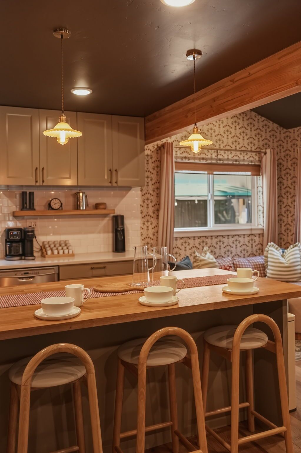 A kitchen with wooden bar stools, a coffee station, and a window with curtains.