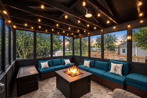 A covered outdoor seating area with a fire feature, large windows, and a view of the outdoors.