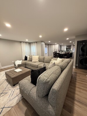 A modern living room with a sectional sofa, ottoman, and a dining area with chairs and a table. - Luxe Home with Michelin Star Kitchen & King Beds (Bolingbrook)