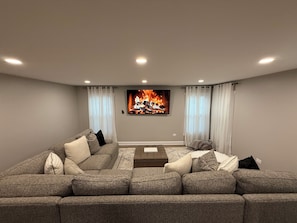 A modern living room with a large sectional sofa, a flat-screen TV mounted above a fireplace, and a neutral color scheme. - Luxe Home with Michelin Star Kitchen & King Beds (Bolingbrook)
