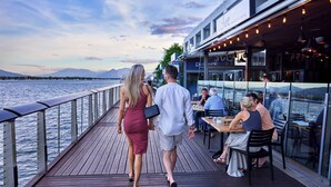 Een houten terras met een balustrade met uitzicht op een waterpartij, een restaurant met terras en bergen op de achtergrond.