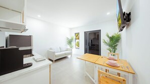 Ein modernes, helles Wohnzimmer mit einem weißen Sofa, einem Holzespeisetisch für zwei Personen und einer Küchenzeile mit Backofen und Mikrowelle.