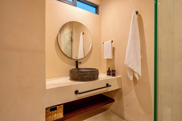 A bathroom with a round mirror, a stone sink, a towel rack, and a small shelf with toiletries. - Loch Collection Tulum (Tulum)