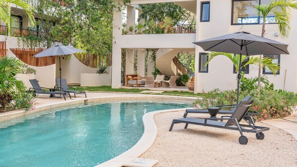 A pool area with lounge chairs and umbrellas, surrounded by greenery and a building with multiple balconies. - Loch Collection Tulum (Tulum)