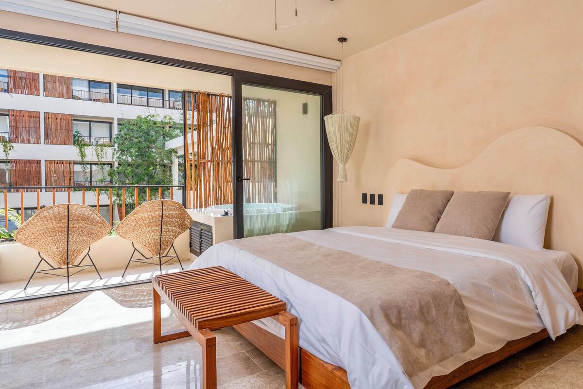 A modern bedroom with a large bed, two bedside tables, a wooden bench, and a view of a building with balconies.