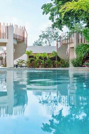Une piscine dont l’eau est si claire qu’elle reflète l’environnement, entourée de palmiers et bordée d’un bâtiment moderne doté d’un escalier en colimaçon.