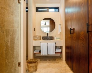 A bathroom with a stone floor, a wooden door, a round mirror, a sink with a stone basin, and a towel rack. - Loch Collection Tulum (Tulum)